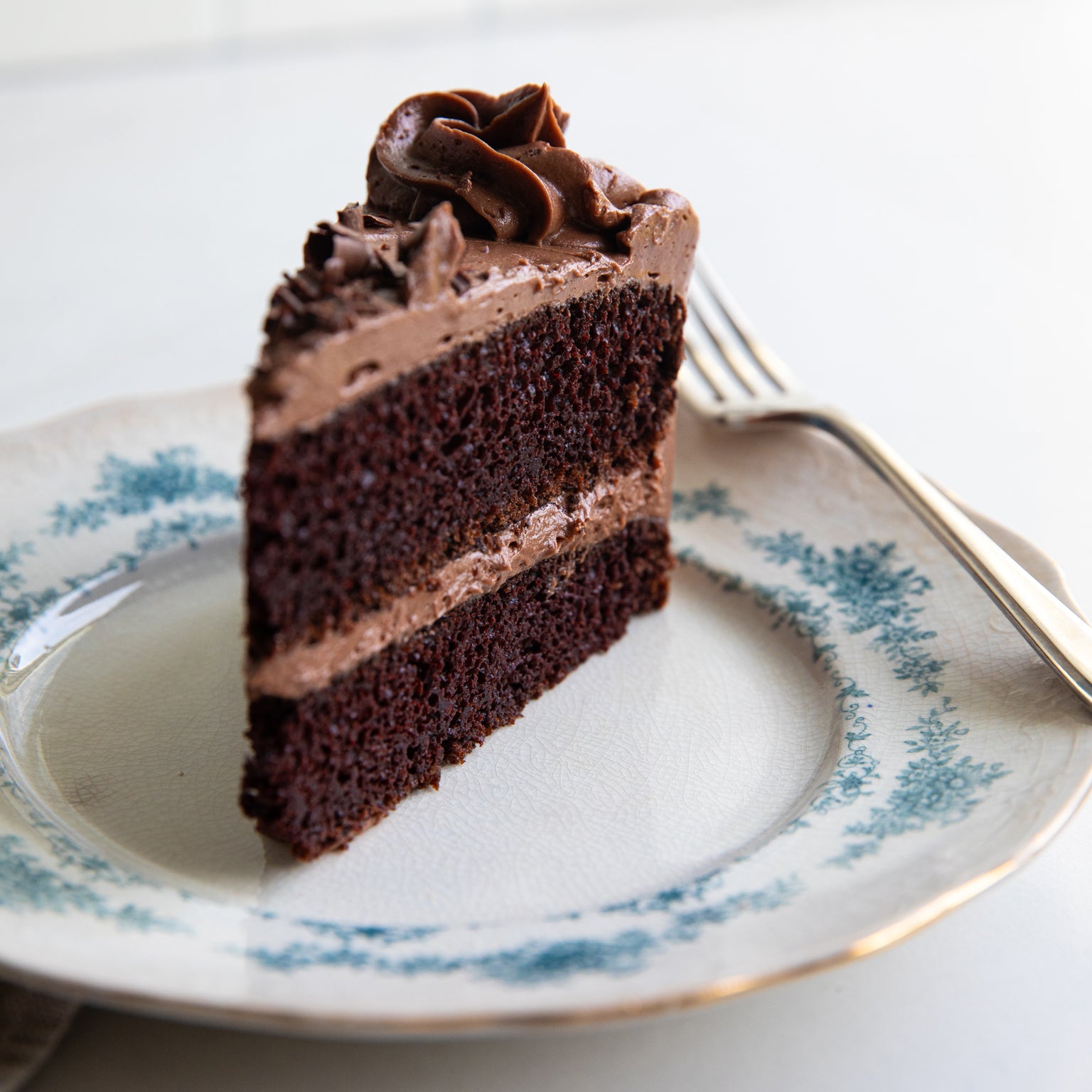 Side view of a slice of Einkorn chocolate cake showing two layers with chocolate buttercream on a white plate with blue floral trim and fork