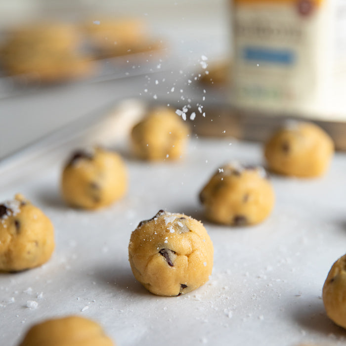 Einkorn chocolate chip cookie dough balls being sprinkled with flaky sea salt on a parchment-lined baking sheet