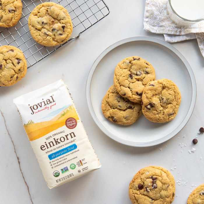 Three einkorn chocolate chip cookies on a plate beside a bag of Jovial einkorn all-purpose flour and a cooling rack