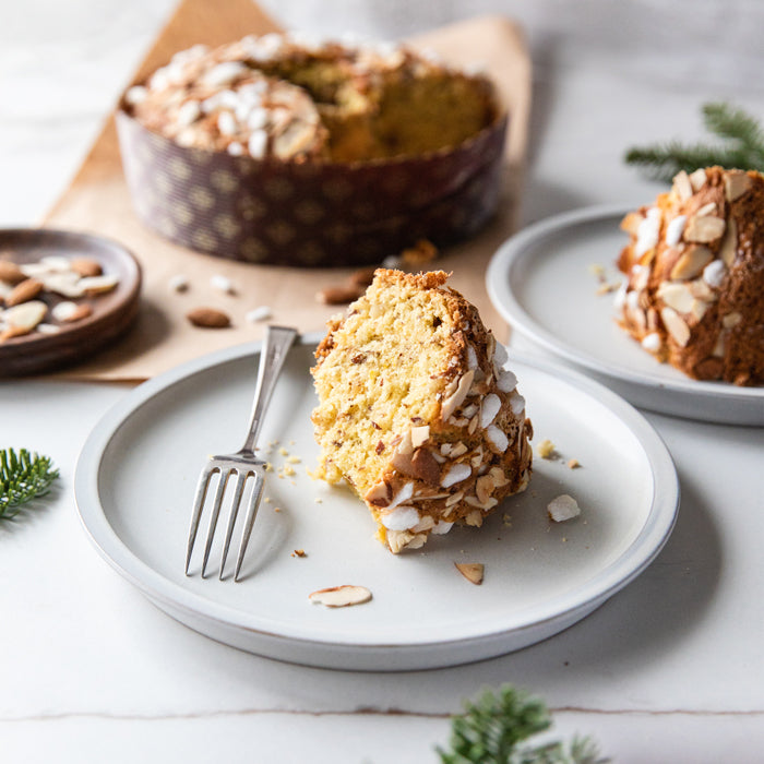 Close-up of a slice of Einkorn Christmas cake topped with sliced almonds on a white plate, whole cake blurred behind