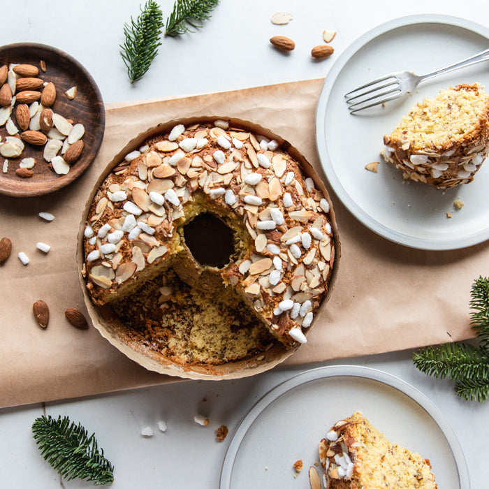 Top-down view of Einkorn Christmas cake topped with sliced almonds and pearl sugar, sliced piece on plate with pine sprigs