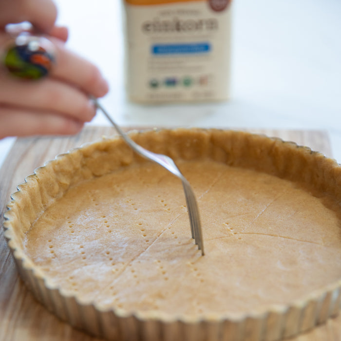 Einkorn tart crust being pricked with a fork in a fluted tart pan on a wooden board, einkorn flour box blurred in the background