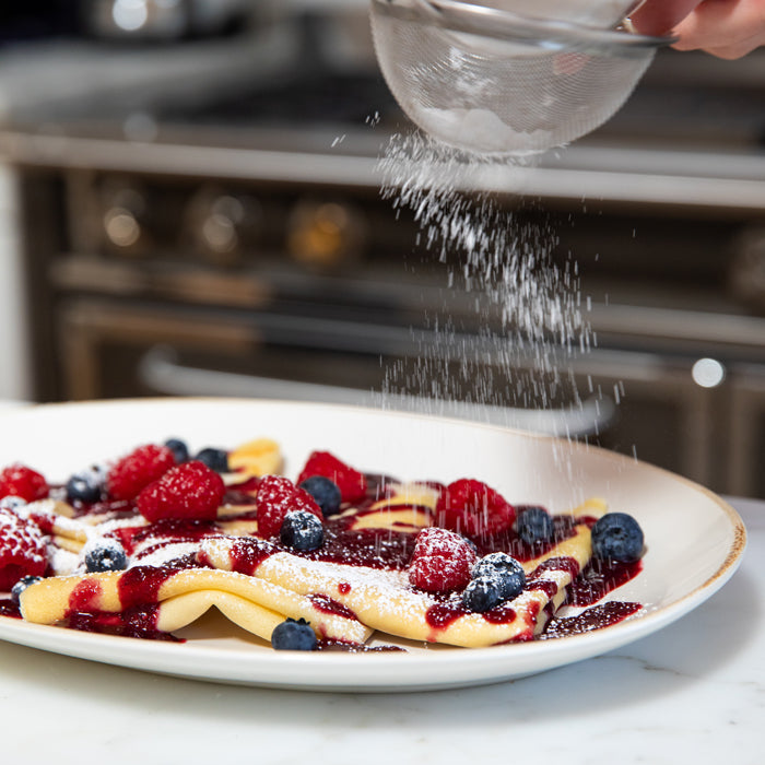 Einkorn crepes with summer berry sauce and berries, being dusted with powdered sugar in a kitchen