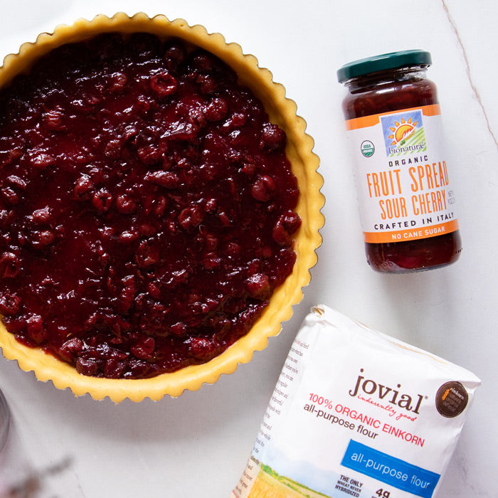 Unbaked tart shell with sour cherry filling beside a jar of sour cherry fruit spread and a bag of Jovial einkorn flour