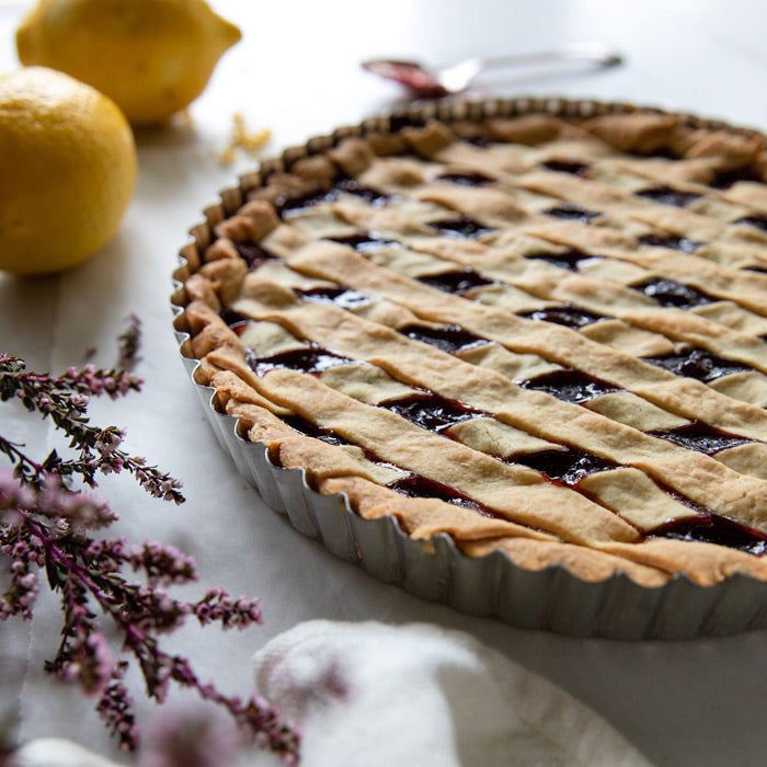 Sour cherry crostata with lattice einkorn crust in a fluted tart pan on a light surface with lemons and flowers