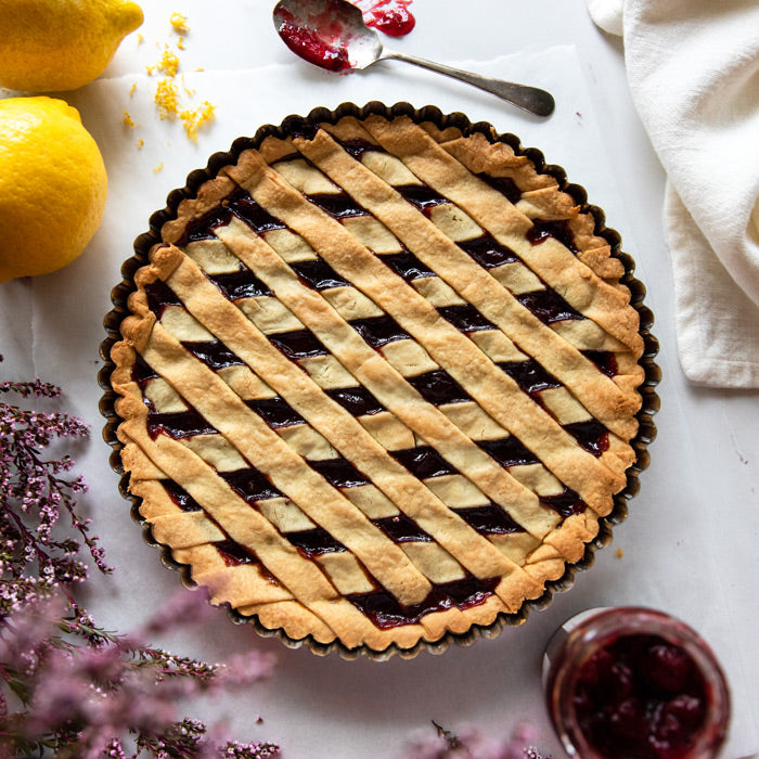 Top-down view of an Einkorn sour cherry crostata with lattice crust in a tart pan, spoon, jar of jam and lemons