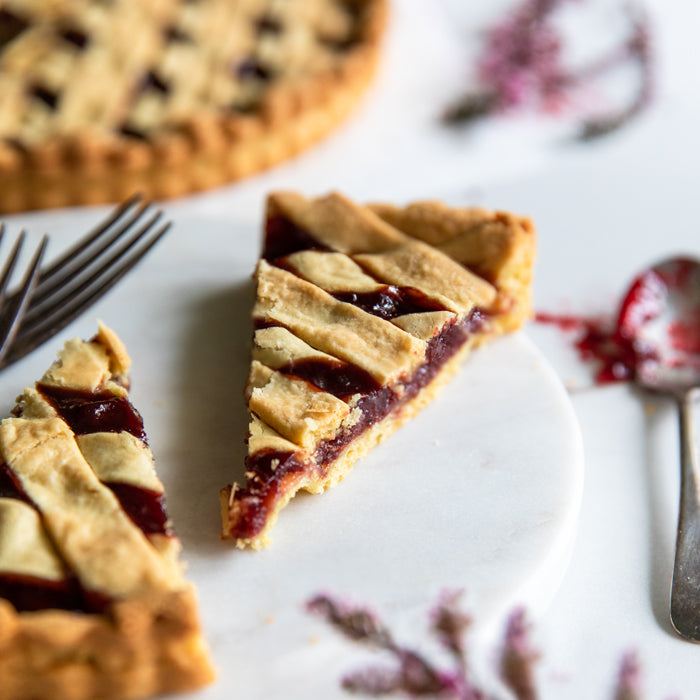 Close-up of a slice of sour cherry Einkorn crostata showing lattice crust and glossy cherry filling on a white marble board