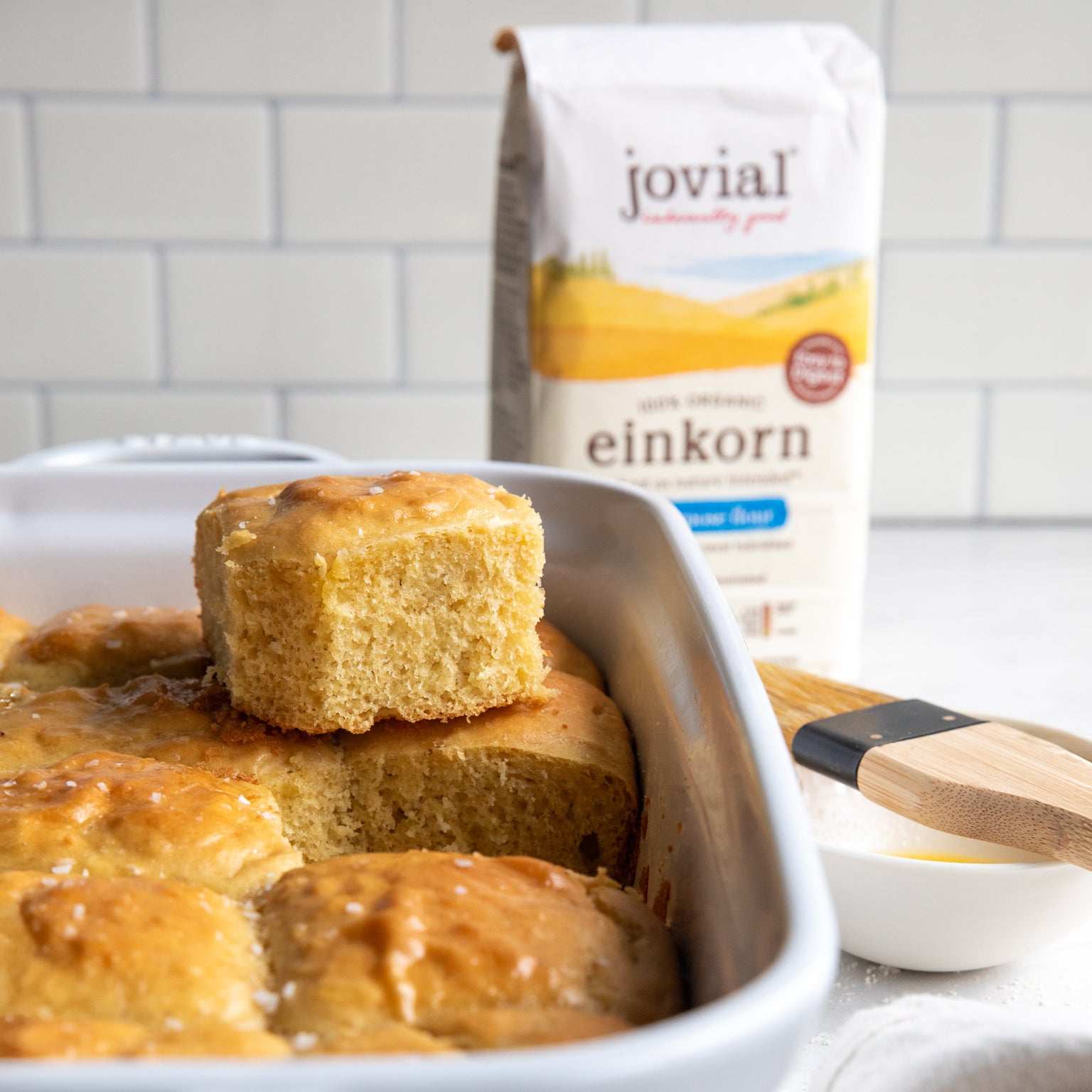 Einkorn dinner rolls baked in a white dish with one roll lifted, Jovial einkorn flour bag and pastry brush in background