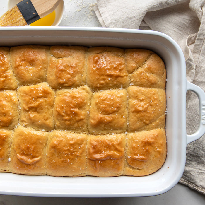Top-down view of a white baking dish with 16 golden-brown einkorn dinner rolls brushed with olive oil on a light linen cloth