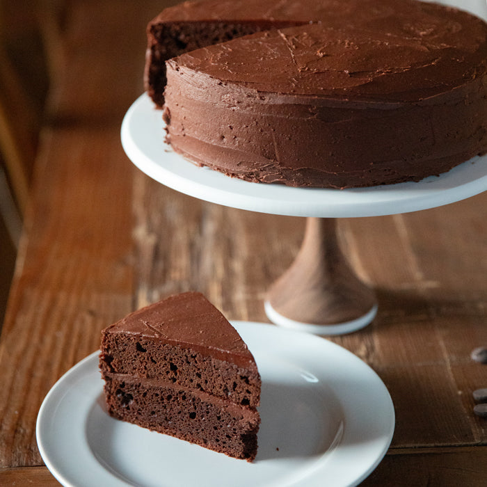 Angled view of a two-layer fudgy mocha Einkorn cake with chocolate buttercream on a cake stand and a plated slice