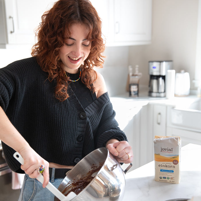 Woman mixing chocolate cake batter in a steel bowl on a kitchen island with a bag of Jovial Einkorn flour visible