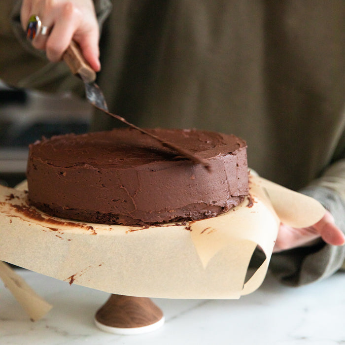 Person spreading chocolate buttercream frosting on a fudgy mocha Einkorn cake on parchment over a wooden cake stand