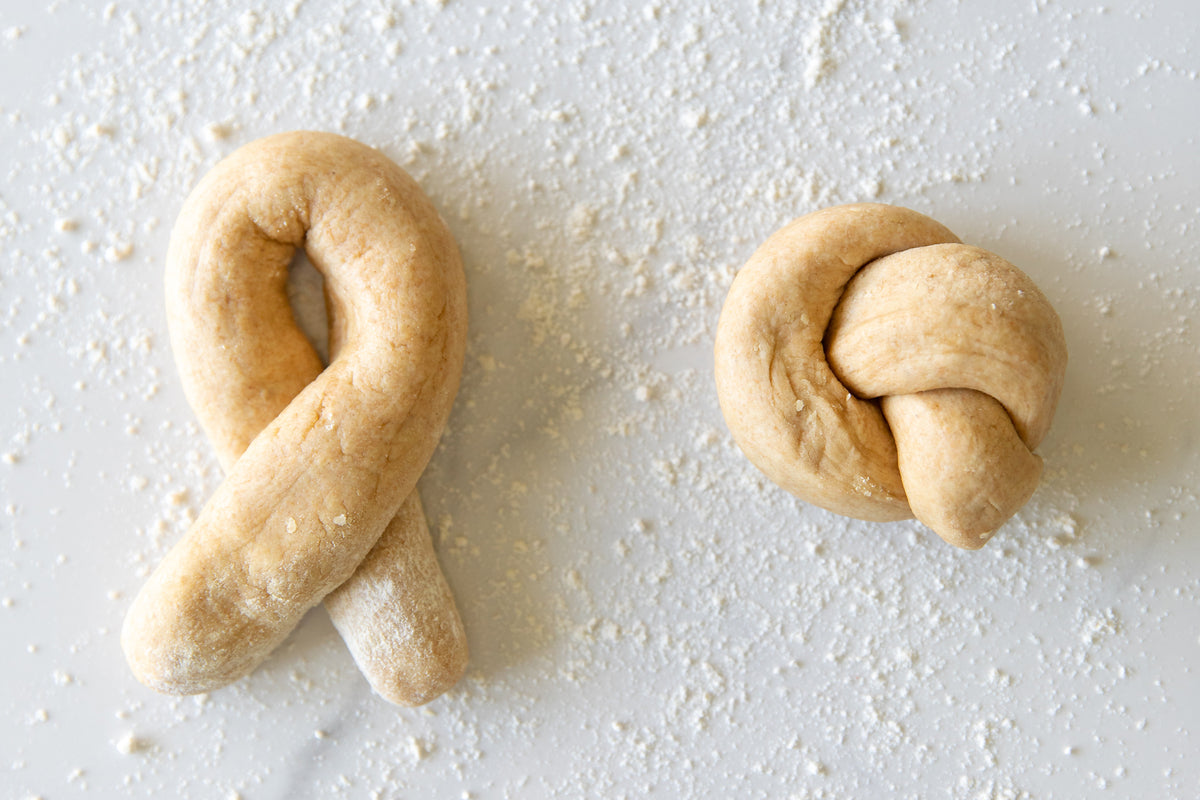Two raw einkorn garlic knot dough pieces on a floured white surface, one twisted and one tied into a knot