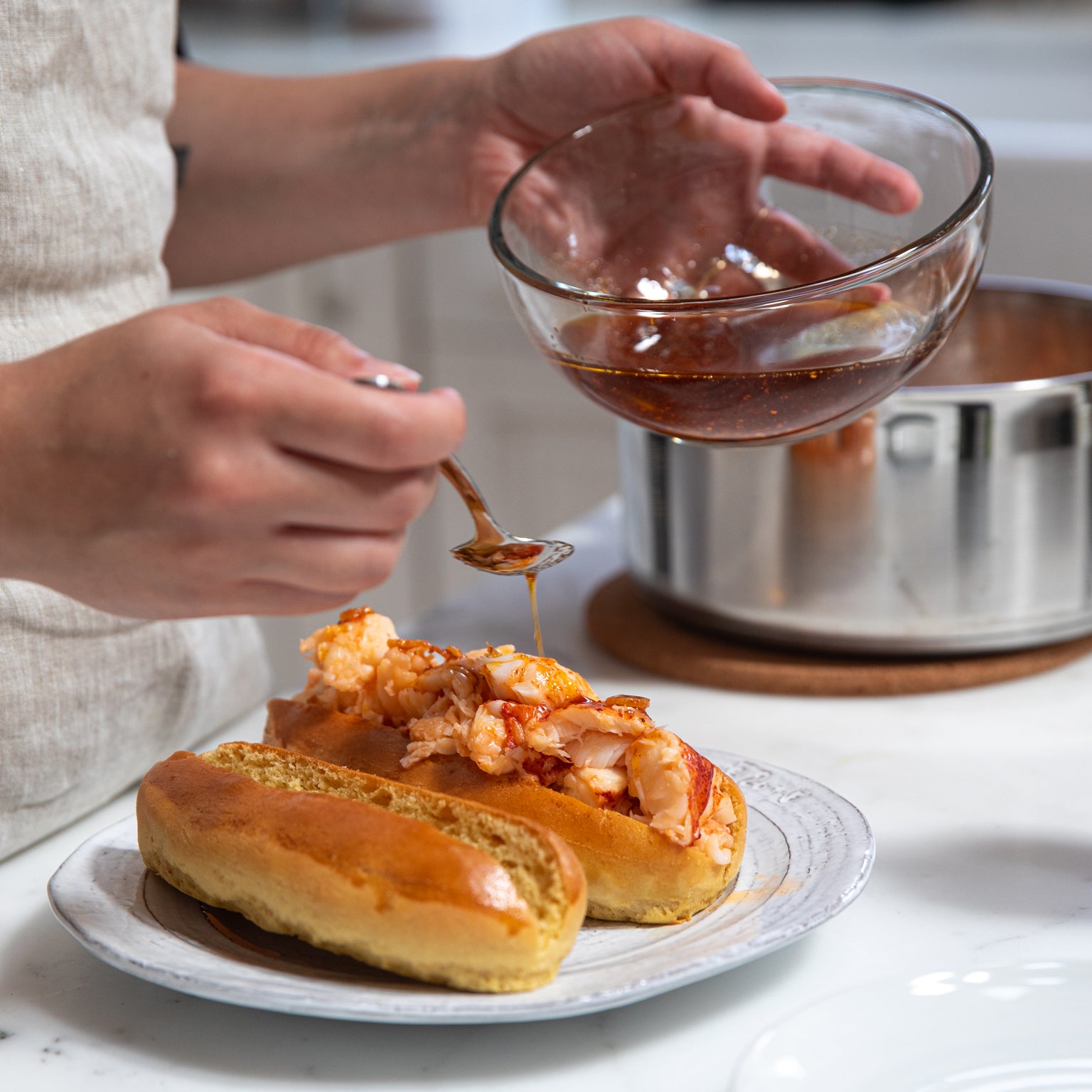 Hands spooning spicy chili oil over a warm lobster roll on a white plate with a second roll and a saucepan in the background