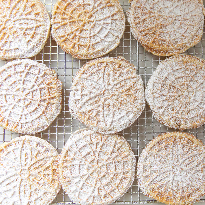 Top-down view of Einkorn pizzelle cookies with pressed decorative pattern, dusted with powdered sugar on a wire cooling rack