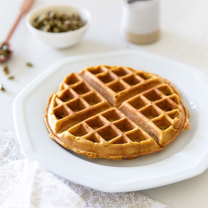 Three-quarter view of a pumpkin pepita einkorn waffle, golden-brown and quartered on a white plate with a bowl of pepitas