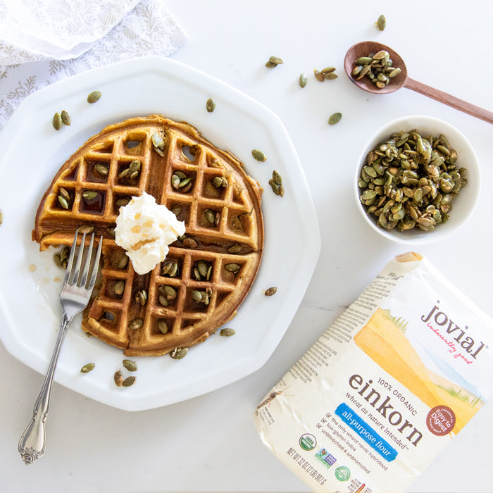 Pumpkin Einkorn waffles topped with whipped cream and candied pepitas on a white plate with a fork and bowl of pepitas nearby