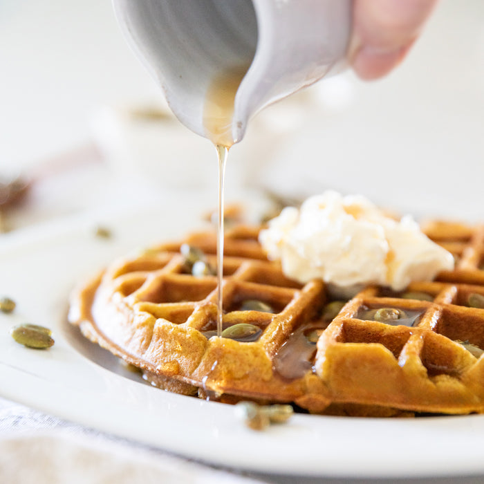 Pumpkin Einkorn waffle topped with candied pepitas and a dollop of butter as maple syrup is poured