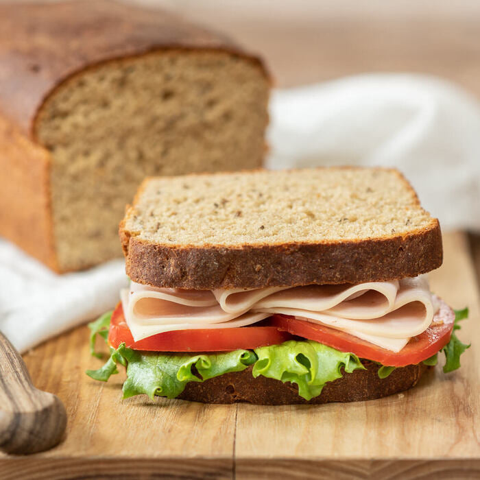 Close-up front view of sliced Einkorn rye bread loaf and a turkey, tomato, and lettuce sandwich on a wooden cutting board