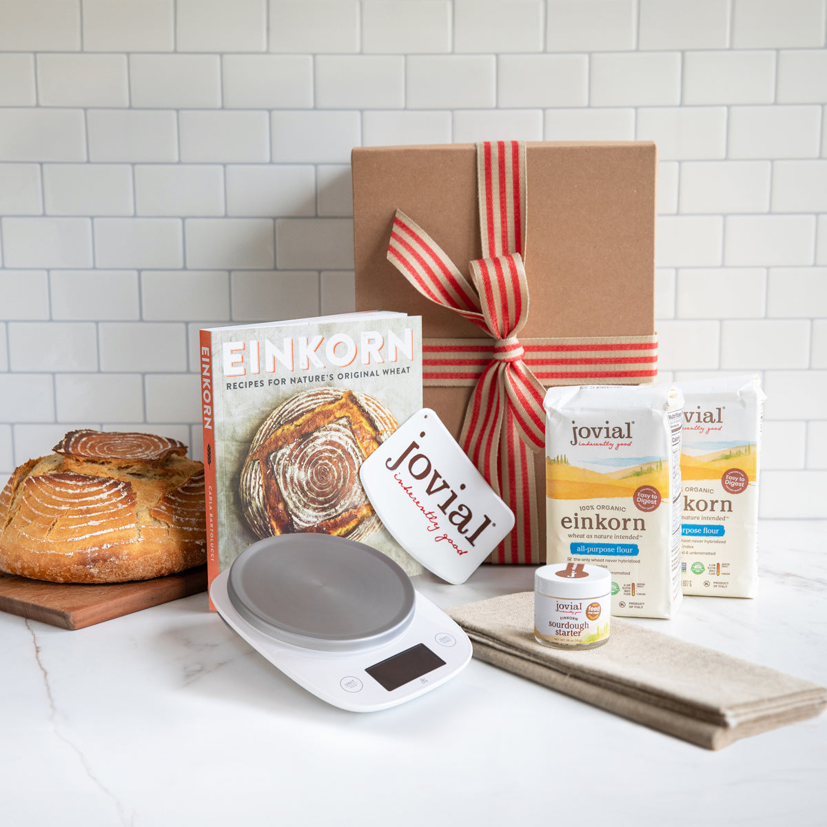 Angled view of Einkorn Sourdough Kit with digital scale, crusty loaf, flour bags, and ribboned gift box on white countertop