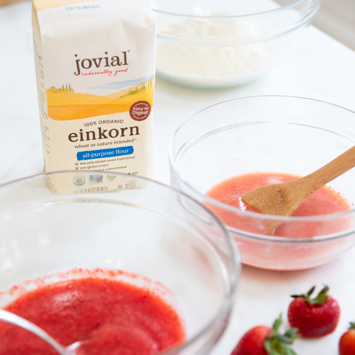 Bowls of strawberry puree with a wooden spoon next to a bag of Jovial Einkorn all-purpose flour and fresh strawberries on a countertop