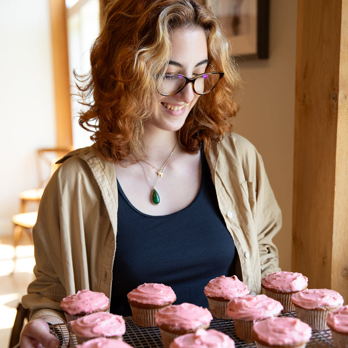 Woman holding a cooling rack of pink strawberry-frosted einkorn cupcakes in a warm kitchen, smiling