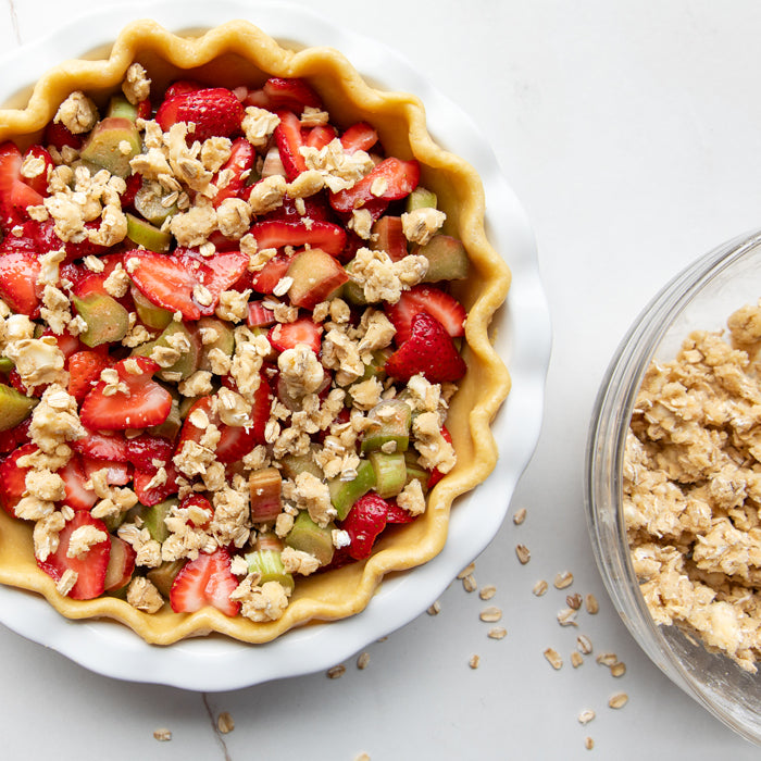 Top-down view of strawberry rhubarb pie with Einkorn crust in a fluted white dish, showing sliced strawberries, rhubarb pieces, and oat crumble topping