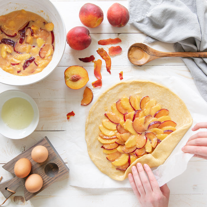Hands folding crust of an Einkorn fruit galette topped with sliced peaches and plums, bowls and eggs on a white surface