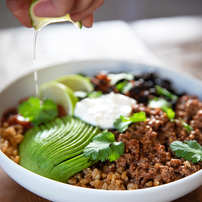 Einkorn wheat berry taco bowl with sliced avocado, seasoned beef, black beans, sour cream, cilantro, and lime being squeezed