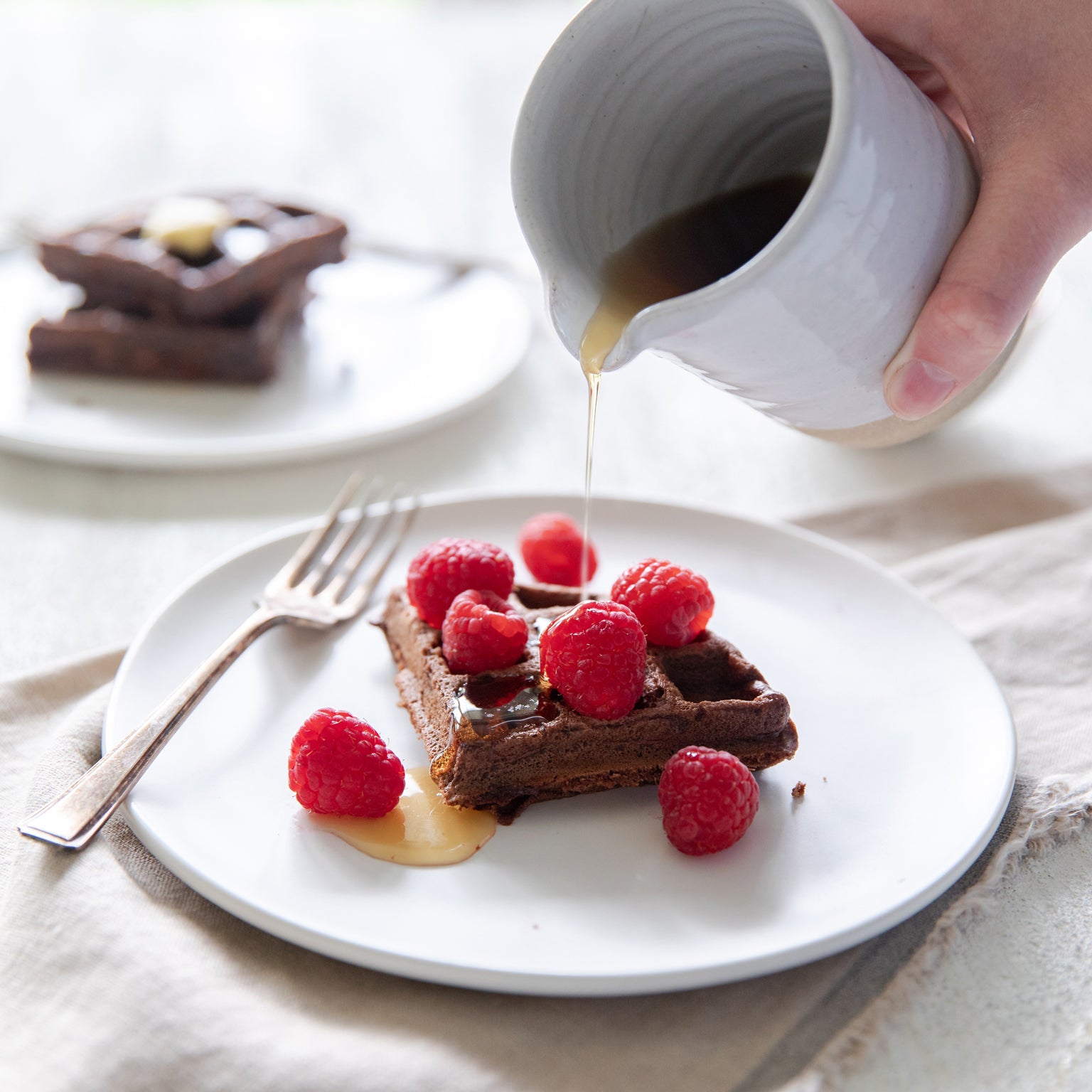Dark chocolate einkorn waffle topped with raspberries as syrup is poured on a white plate
