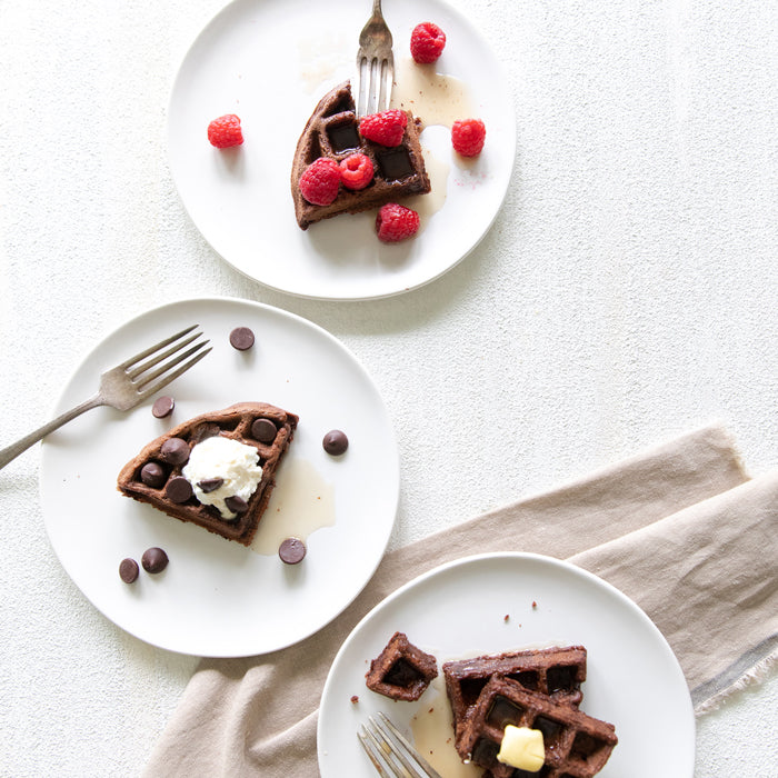 Three plates of dark chocolate einkorn waffle slices topped with raspberries, chocolate chips, and butter