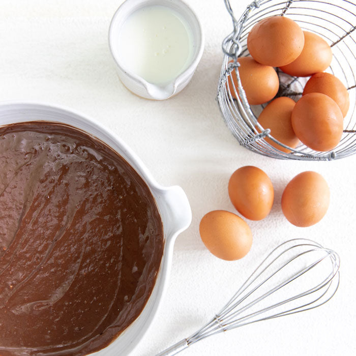 Dark chocolate einkorn waffle batter with eggs, whisk, and milk pitcher on white background