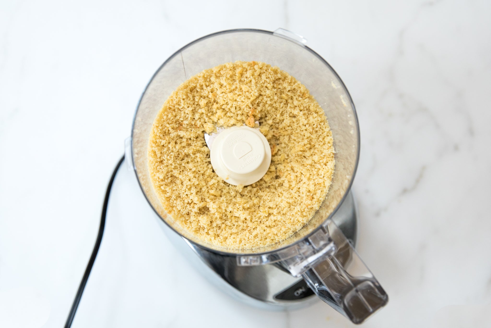 Food processor containing homemade Einkorn breadcrumbs on a white marble countertop
