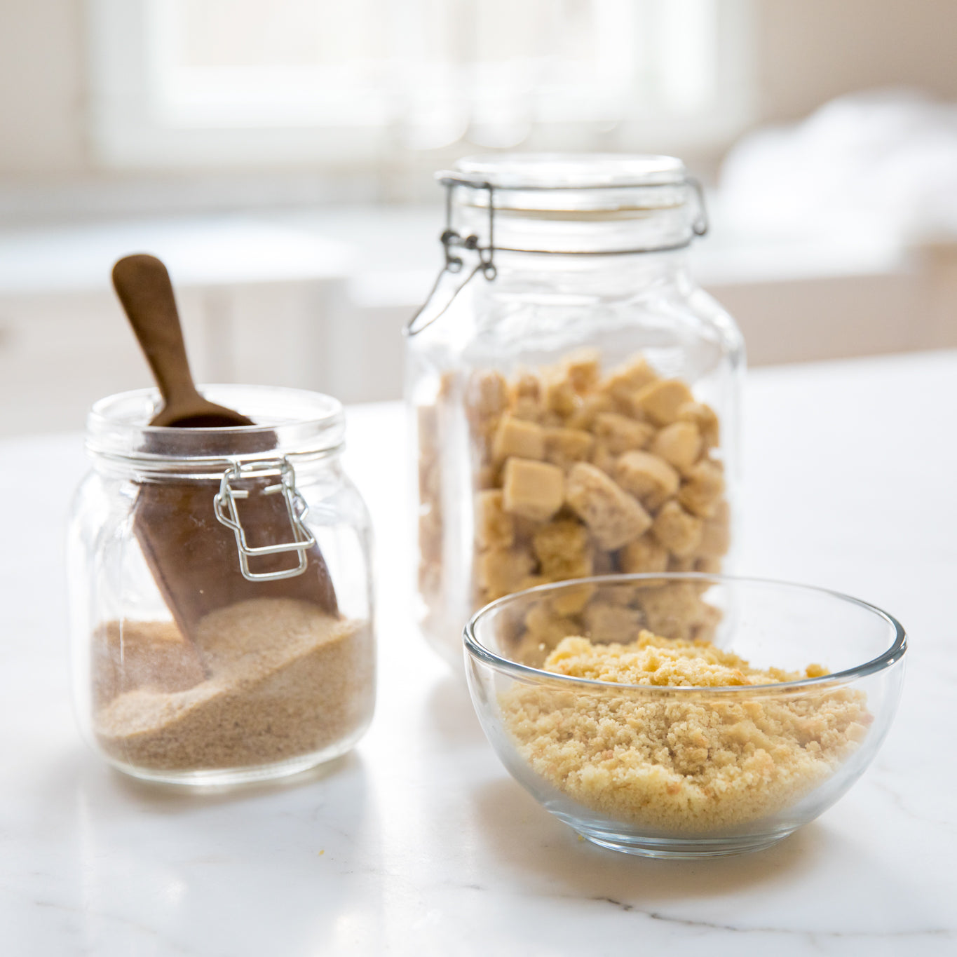 Front view of Einkorn breadcrumbs in a glass bowl showing fine texture with jars of bread cubes on a marble countertop