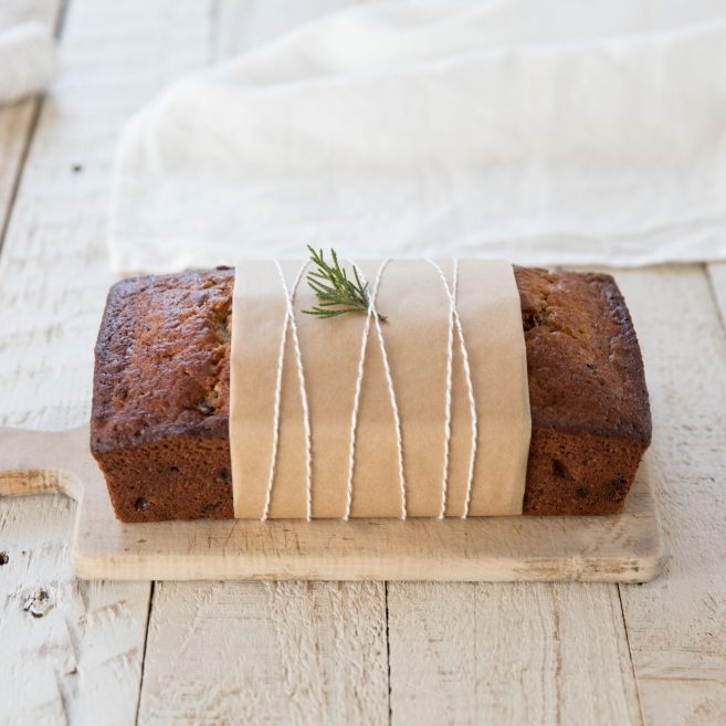 Front view of Einkorn rum fruit cake wrapped in kraft paper and twine, topped with a rosemary sprig on a wooden cutting board