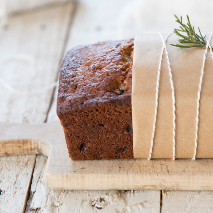 Three-quarter view of honey-sweetened rum fruit cake loaf wrapped in kraft paper and tied with twine on a rustic wooden cutting board