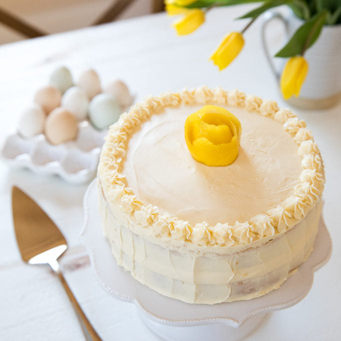 Three-quarter view of Einkorn lemon layer cake with creamy frosting and lemon-rose topper on a white stand, eggs in background