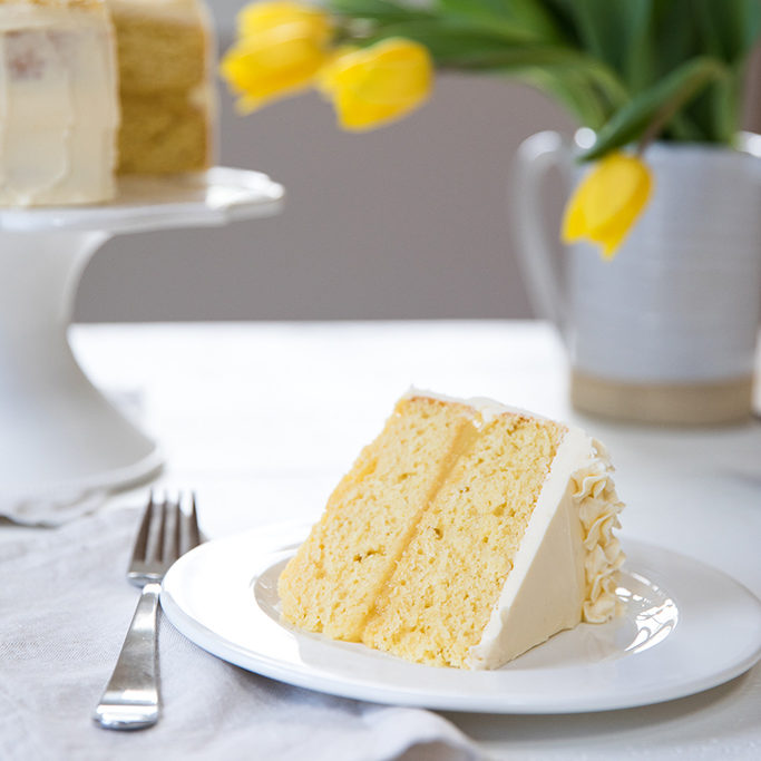 Slice of Einkorn lemon layer cake with white buttercream on a plate, blurred cake and yellow tulips in background