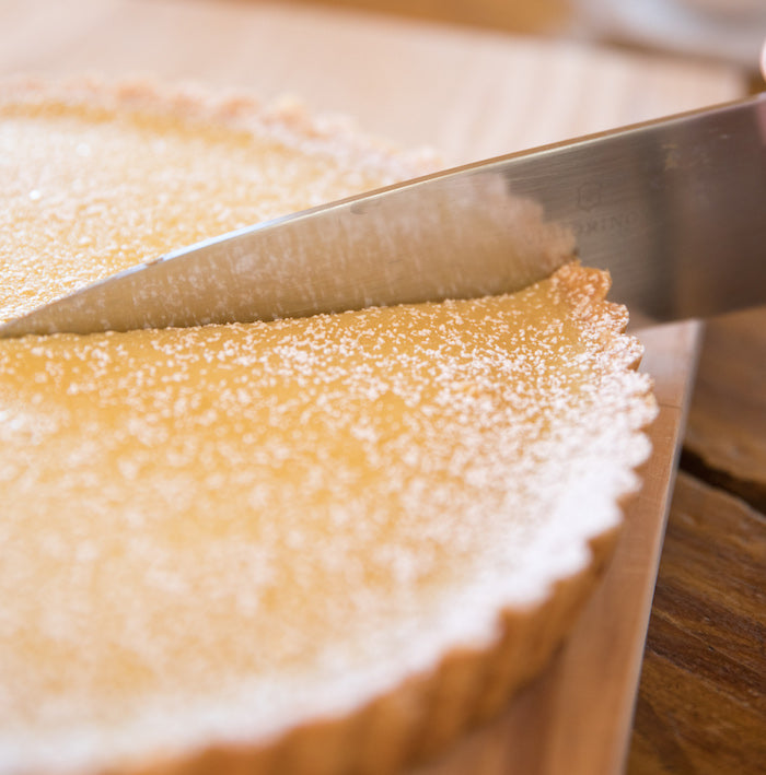 Close-up of Einkorn lemon cream tart being sliced, smooth lemon filling dusted with sugar and fluted crust on wood board