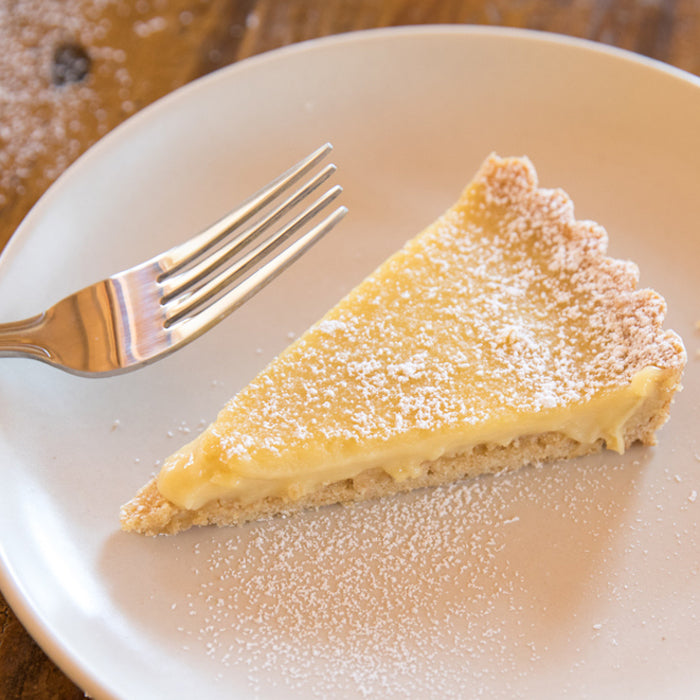 Three-quarter top view of an Einkorn lemon cream tart slice on a white plate dusted with powdered sugar, fork beside it