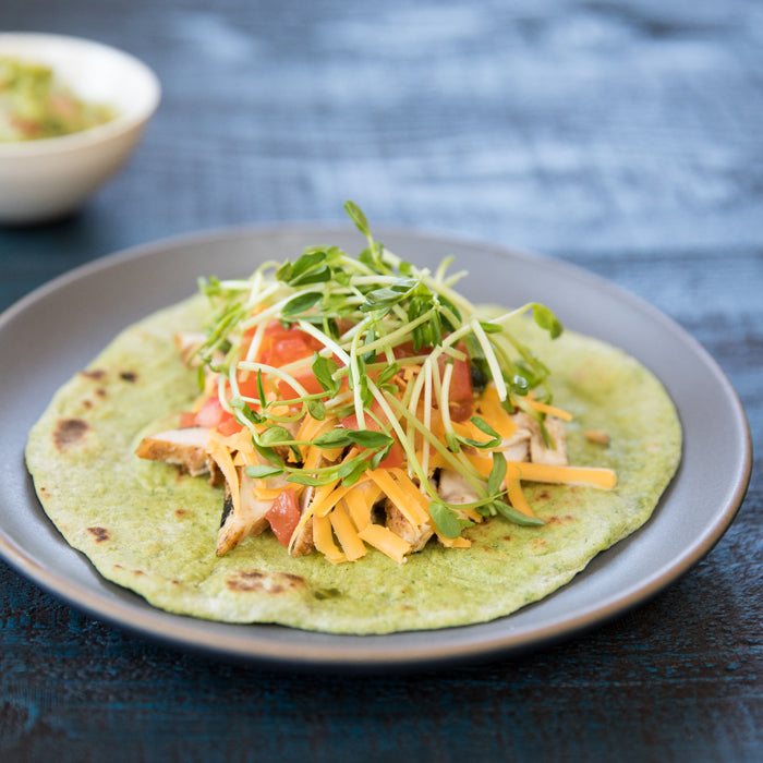 Top-down view of an Einkorn spinach tortilla topped with chicken, cheddar, tomato and microgreens on a gray plate