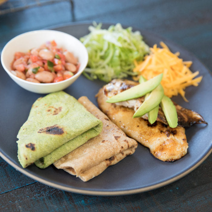 Spinach and whole wheat einkorn tortillas served on a plate with grilled fish, avocado slices, shredded lettuce, cheddar, and a bowl of beans