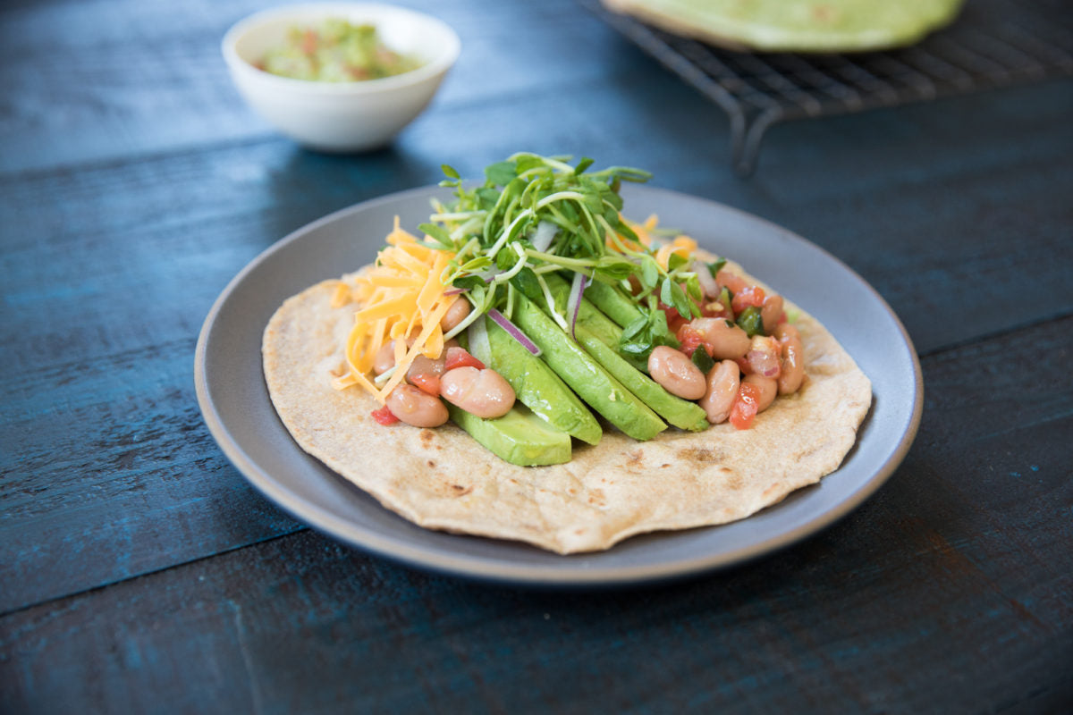 Open whole wheat einkorn tortilla topped with sliced avocado, white beans, shredded cheddar and microgreens on a gray plate