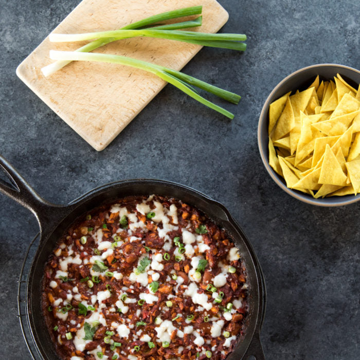 Skillet of chicken enchilada bean dip topped with melted cheese and sliced green onions, bowl of tortilla chips nearby