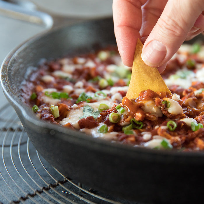 Hand dipping an olive oil chip into a cast-iron skillet of cheesy enchilada-style dip topped with sliced green onions