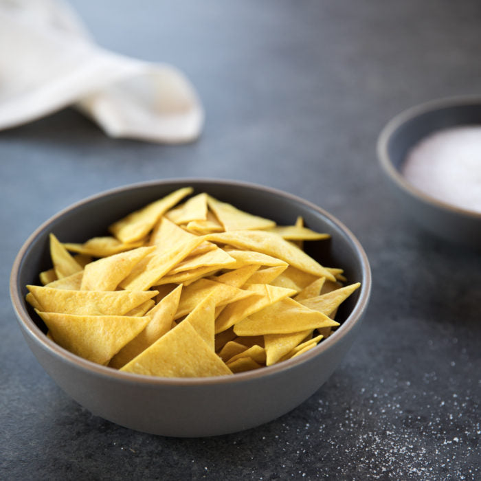 Overhead view of a gray bowl filled with golden triangular olive oil chips on a dark stone surface with scattered salt