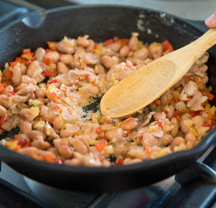 Chicken and pinto beans with diced red pepper and onion being stirred in a cast-iron skillet on a stovetop