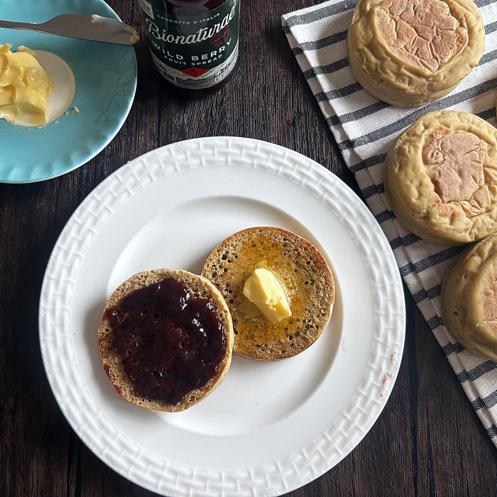 Overhead view of sourdough Einkorn English muffins showing a split toasted muffin with butter and berry jam on a white plate