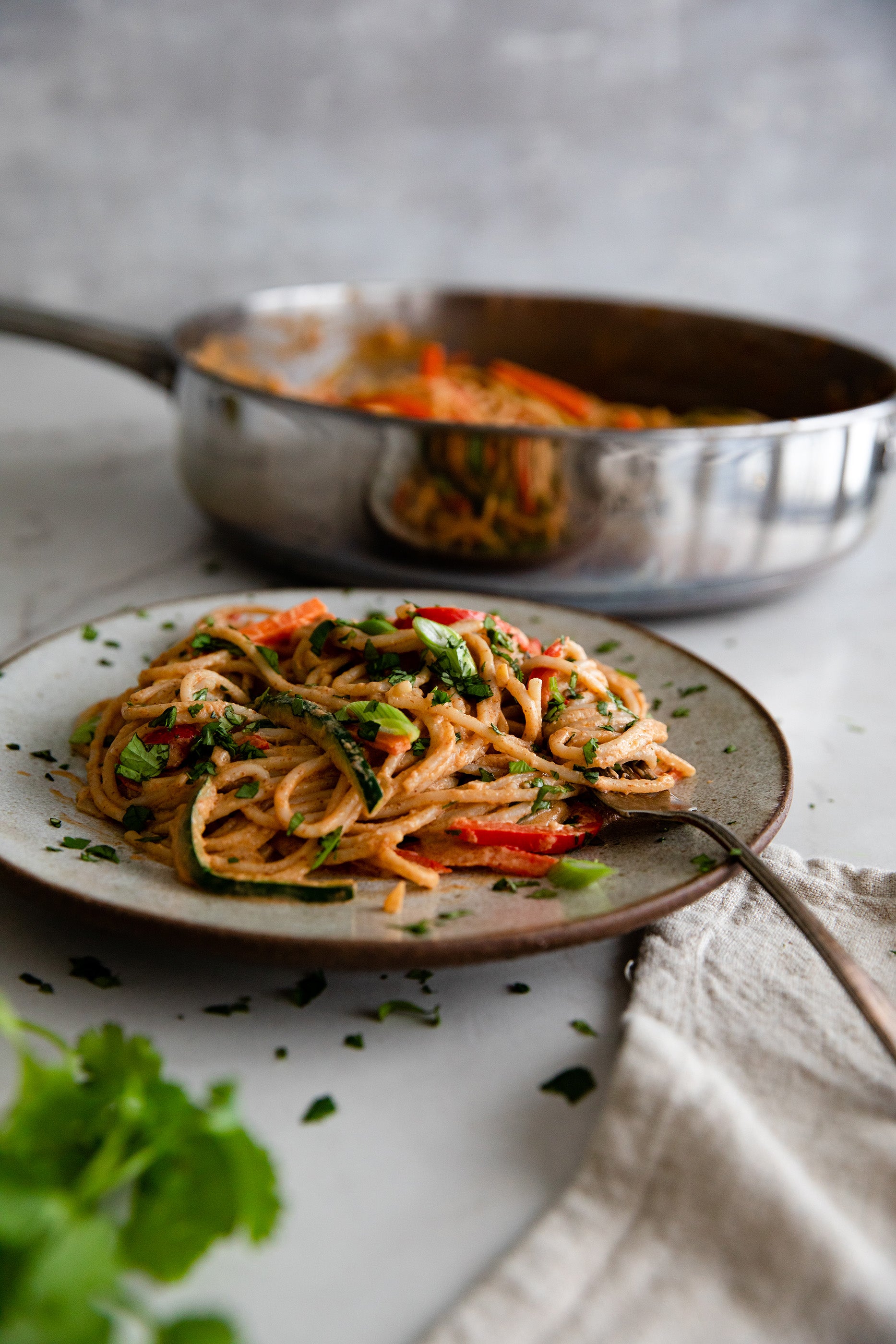 Side view of brown rice spaghetti in peanut sauce with zucchini and red pepper, plate with skillet behind