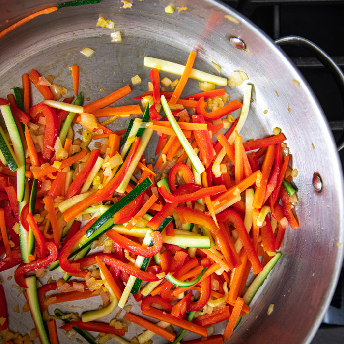 Julienned summer vegetables red pepper carrot zucchini and onion sautéing in a stainless-steel skillet for spaghetti with peanut sauce